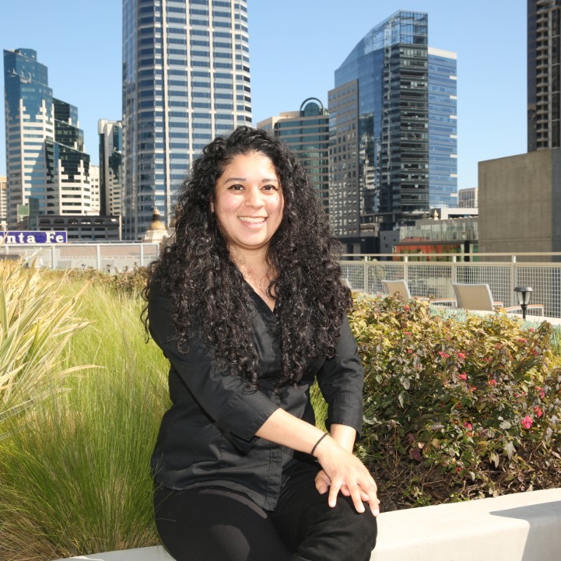 A smiling Jessica Ramos-Bahena, wearing a black shirt and pants, is sitting outside in front of a scenic downtown San Diego background.