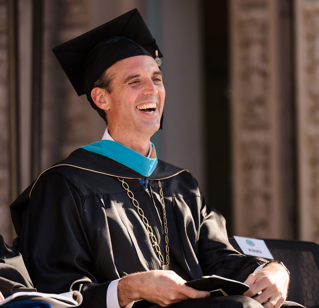 Greg Smith wears a black graduation cap and gown with a teal sash and a chancellor medallion