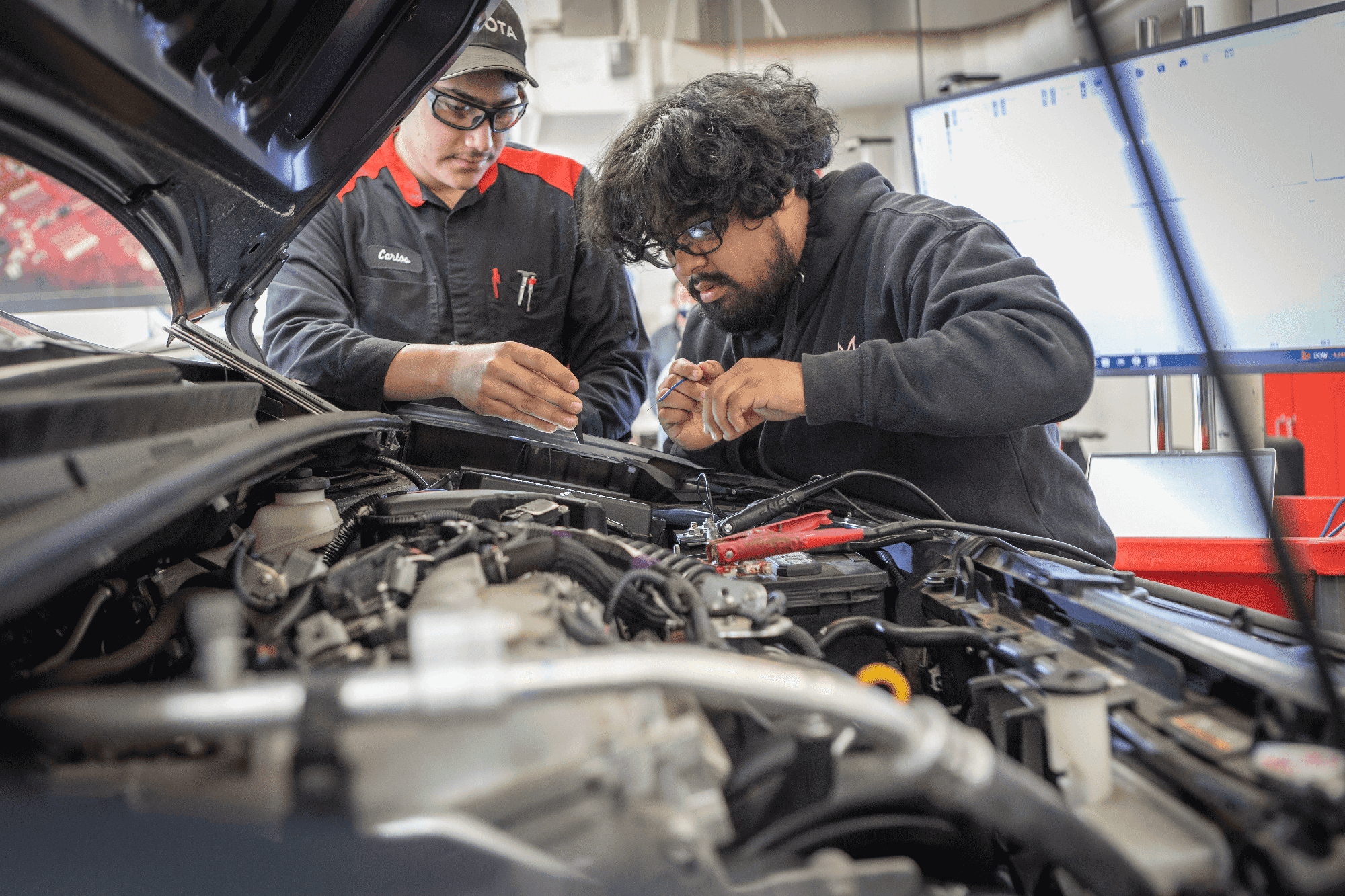 A student is working on a car engine in an automotive class. He is wearing safety goggles and the hood of the car is propped up over his head