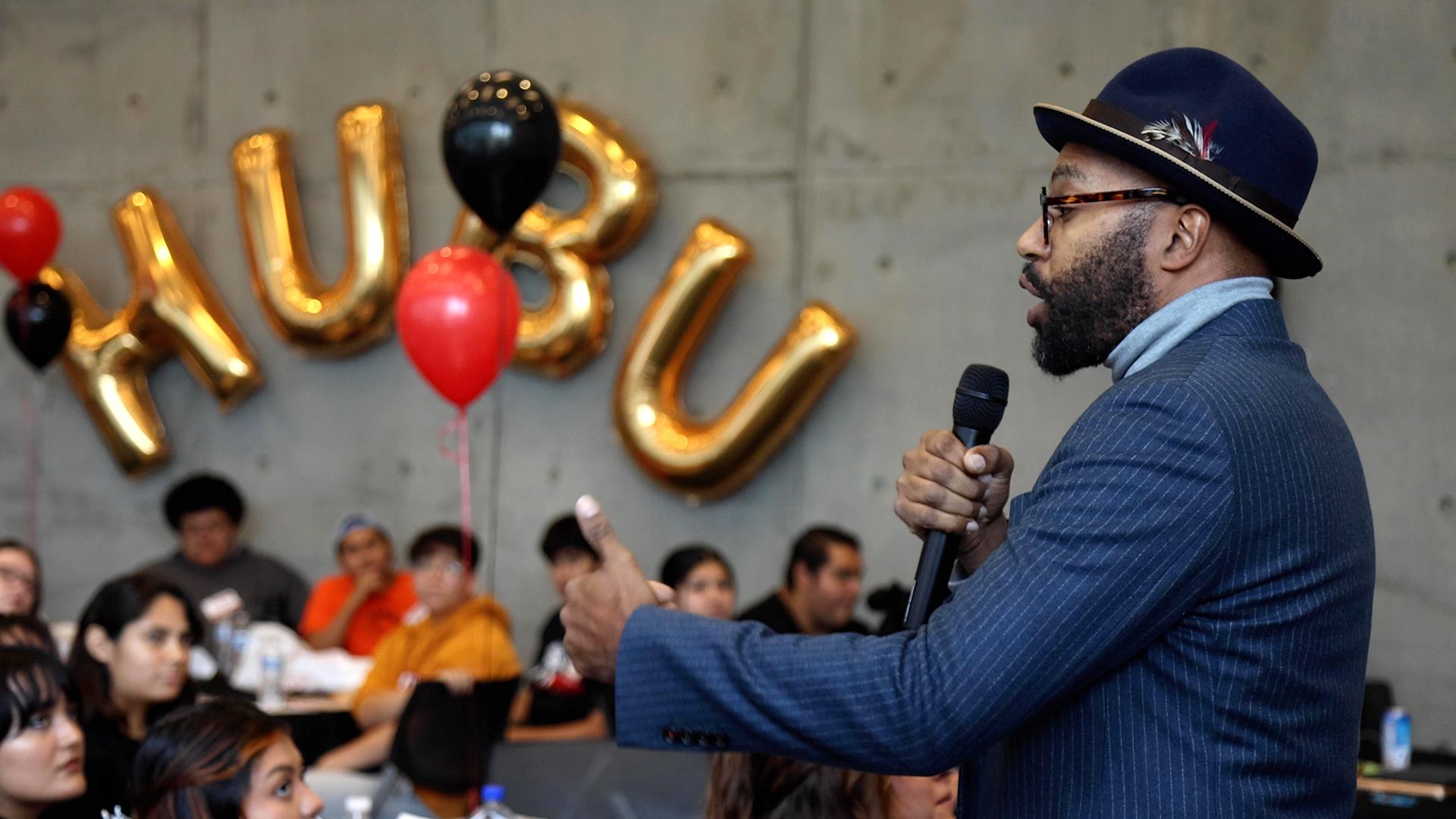 A speaker at a hubu conference speaks to people sitting at tables. There are gold balloons on the wall that spell HUBU