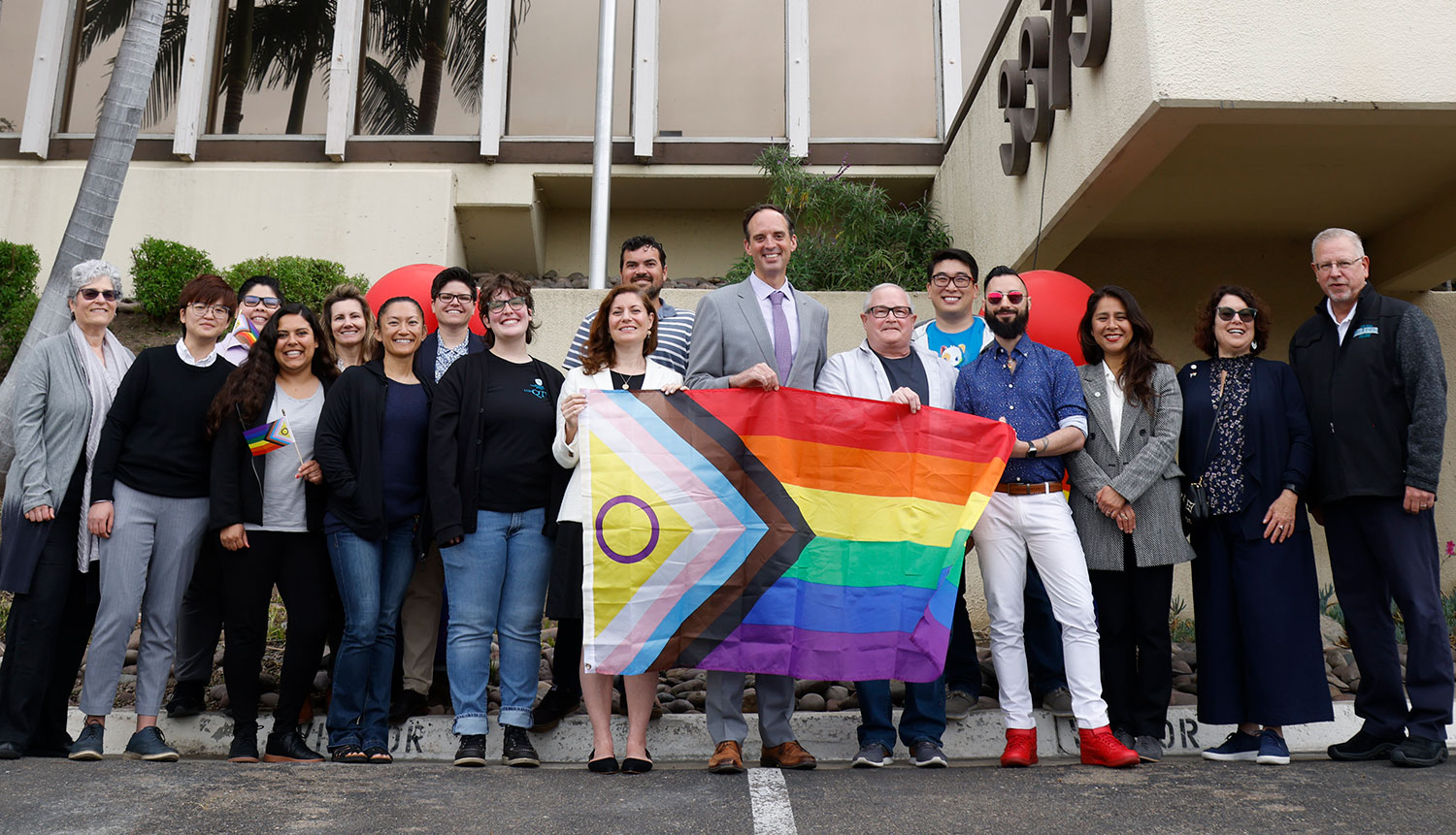 17 people in a group photo pose with a pride flag before the flag is raised for the first time at the district office.