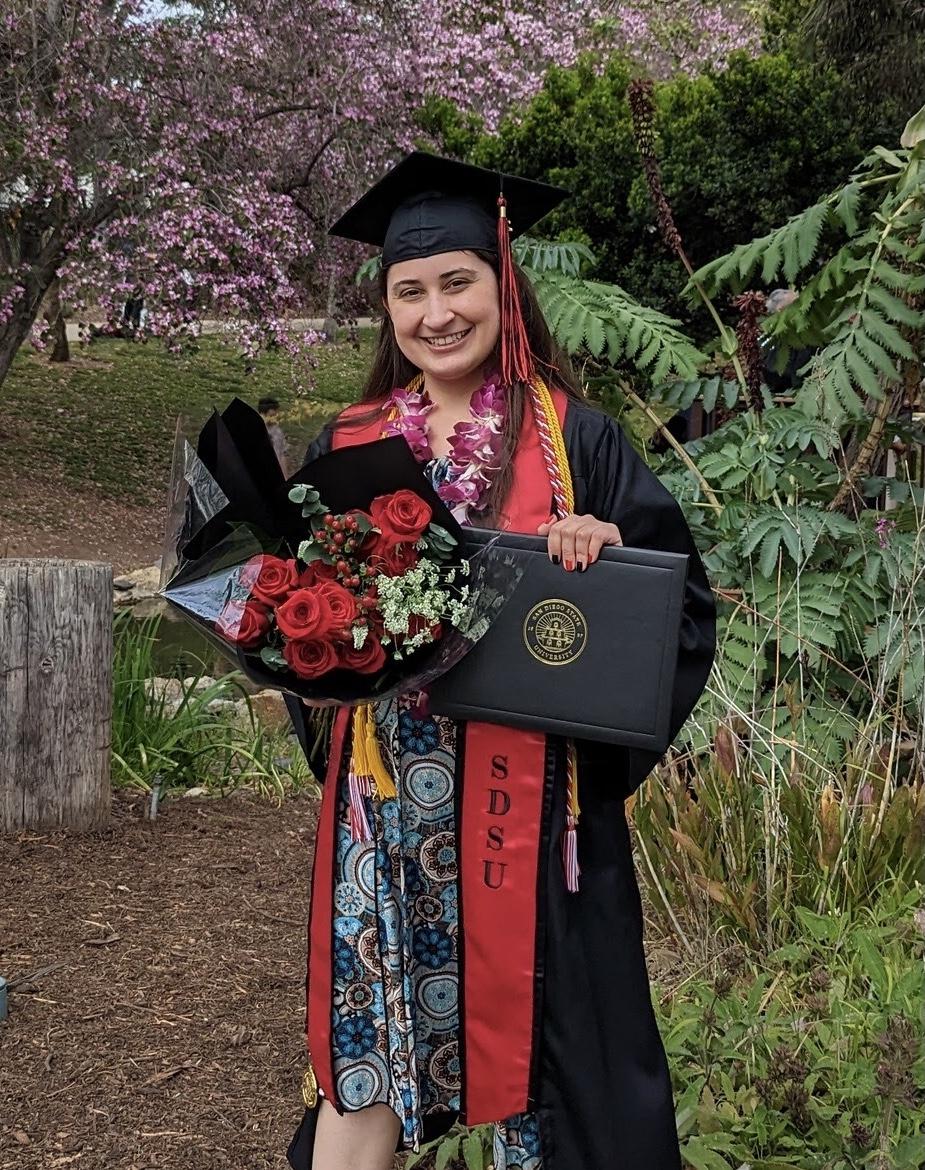 Cassandra is in her graduation regalia holding a boquet of flowers and diploma smiling