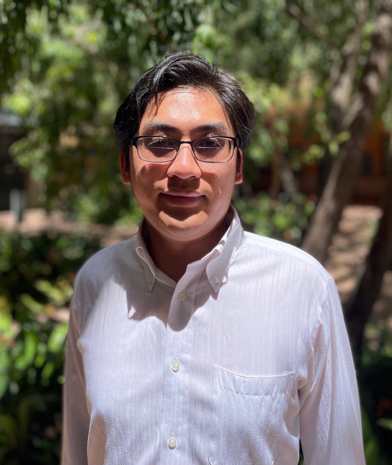 Luc Huynh is standing outside on Miramar campus, with trees behind him. He’s wearing a white shirt and glasses and looking at the camera.