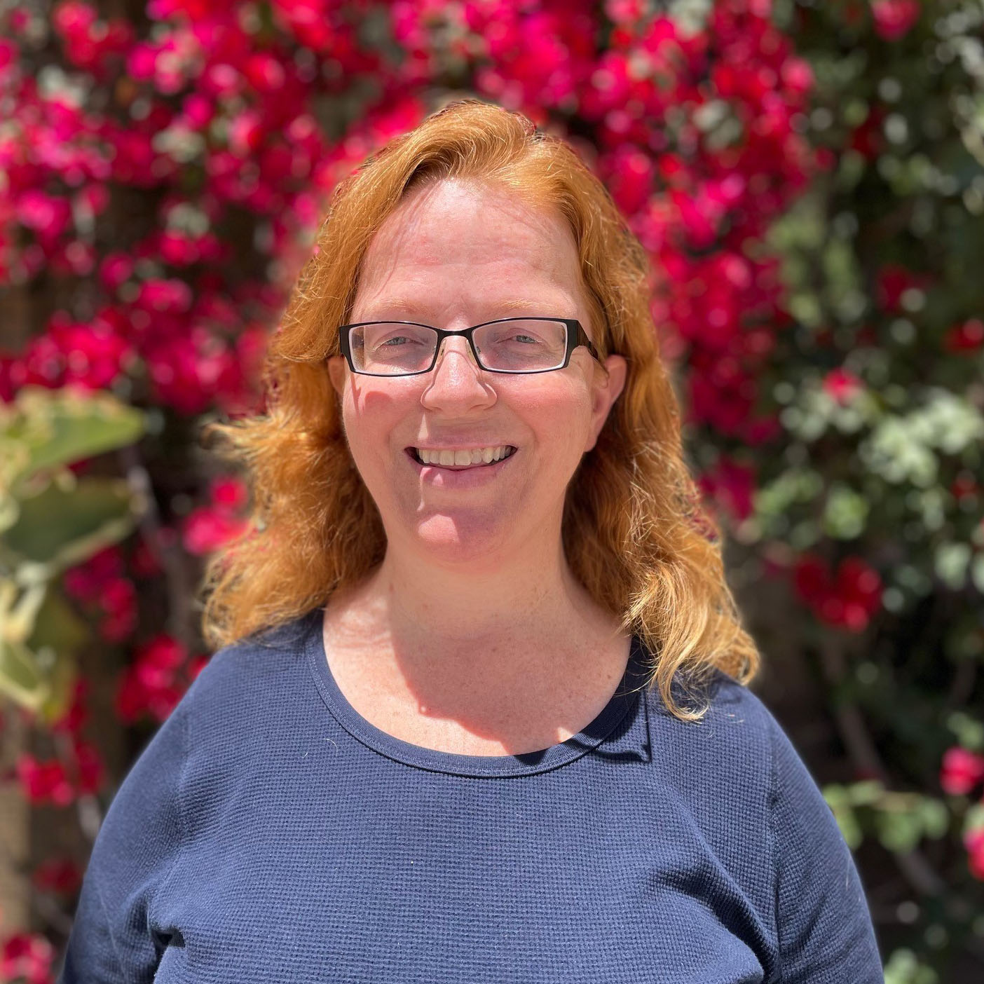 Margaret Reed is standing outside on Miramar campus, with red flowering foliage behind her. She’s wearing a blue shirt and glasses and smiling at the camera.