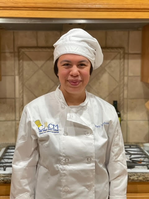 Mary Mendoza is standing in a kitchen and smiling. She is wearing a white chef’s hat and coat.