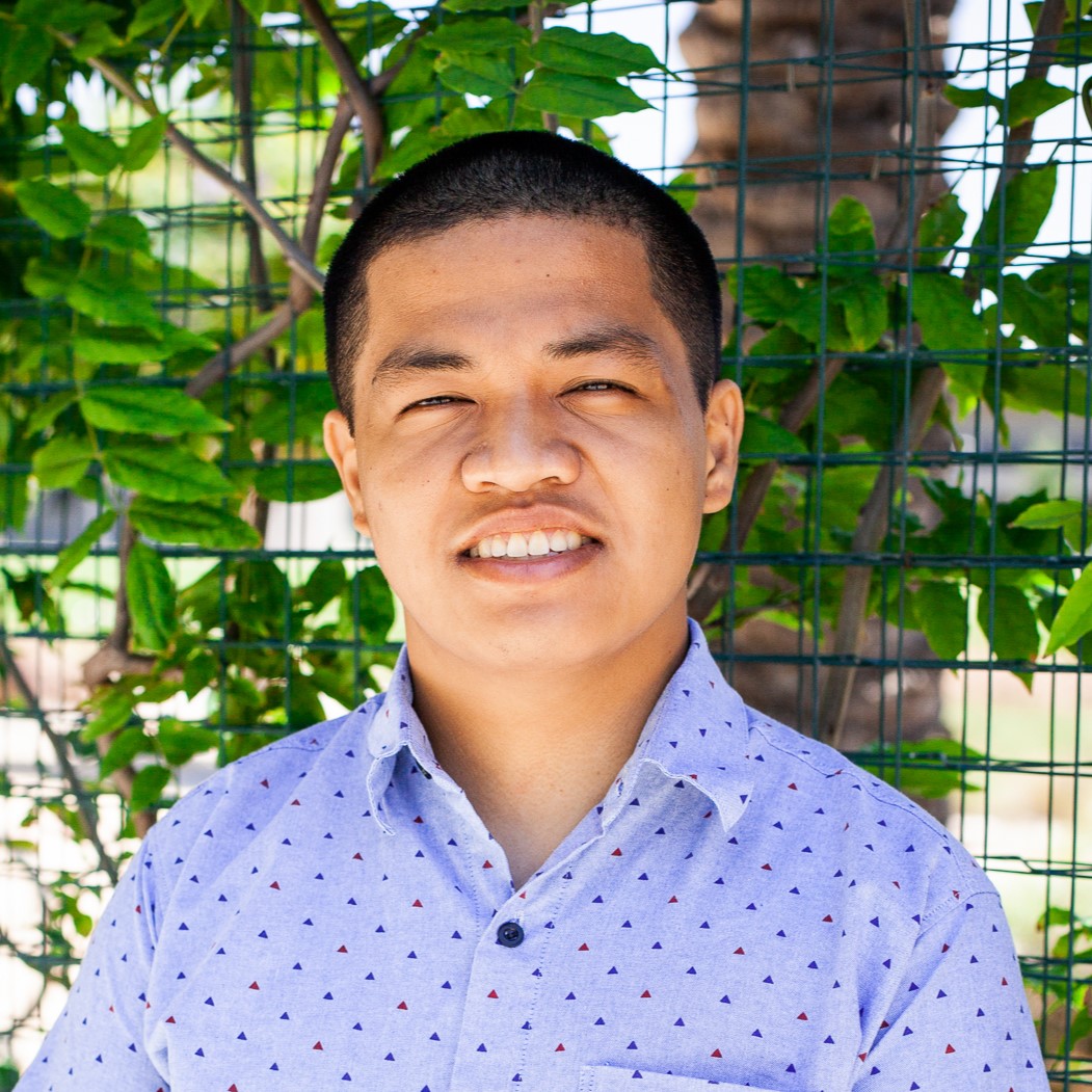 Michael Sarmiento is smiling, standing in front of fencing that is covered in green foliage. He’s wearing a light blue button-down shirt with red and navy blue accents.