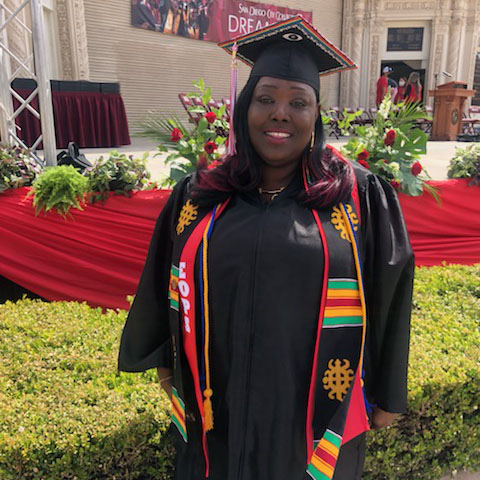 Raynisha Jackson, dressed in graduation regalia, stands smiling in front of City College commencement stage at Spreckels Organ Pavilion in Balboa Park.