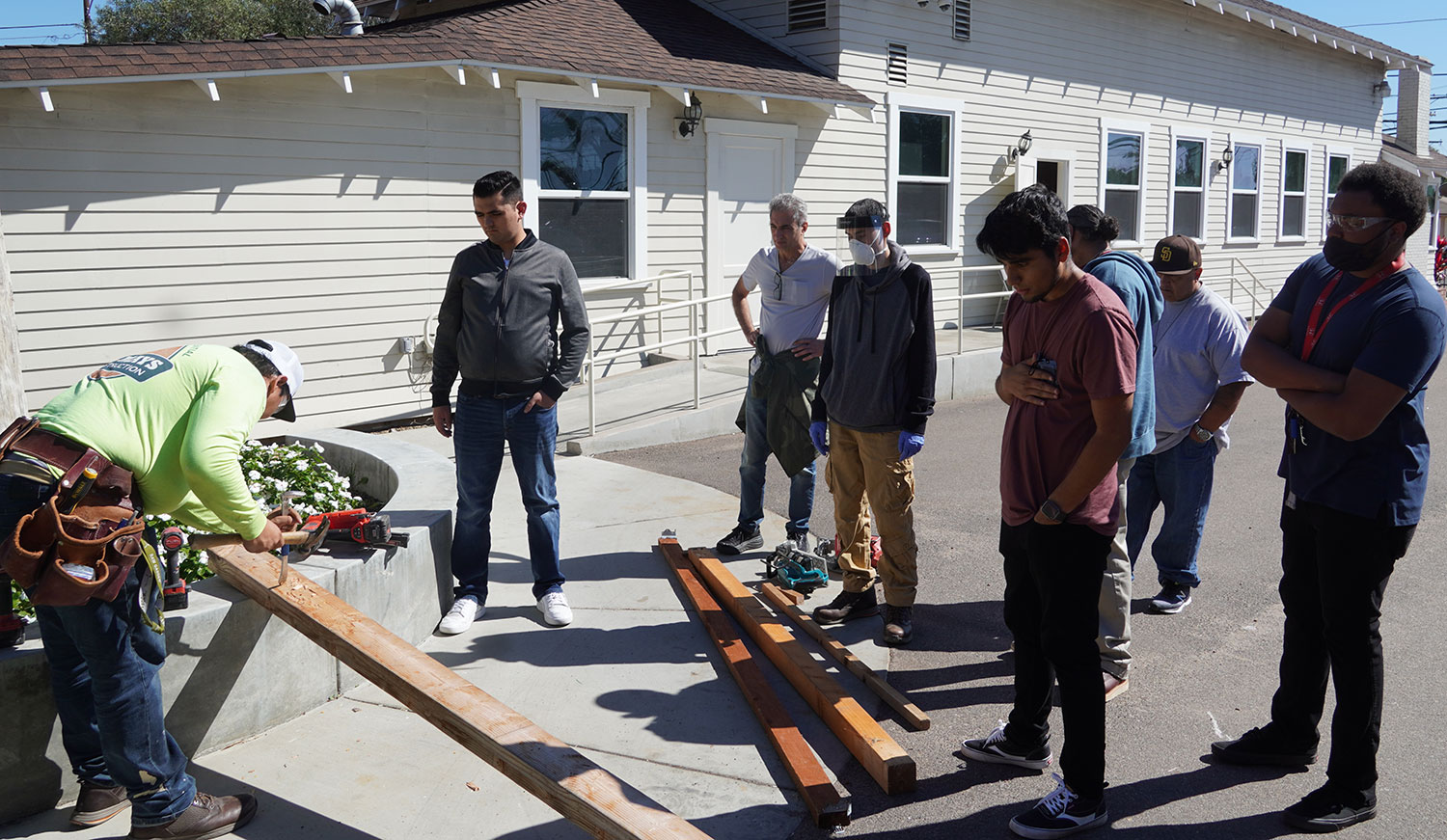 Seven students watch as another student uses a hammer and a chisel on a piece of wood