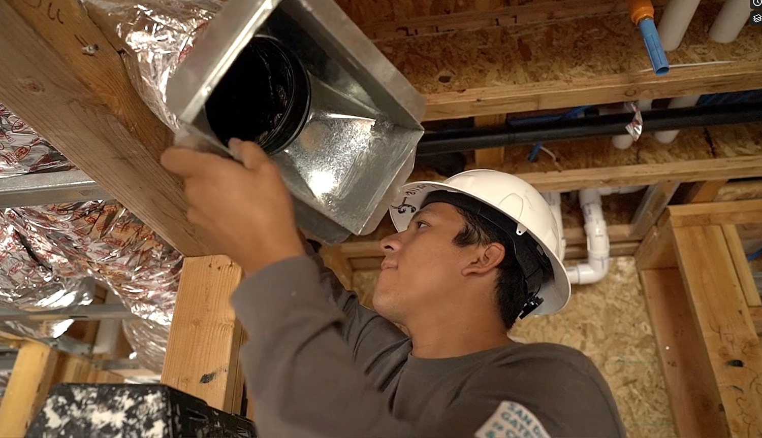 A student in a white hard hat climbs a ladder to reach an air duct