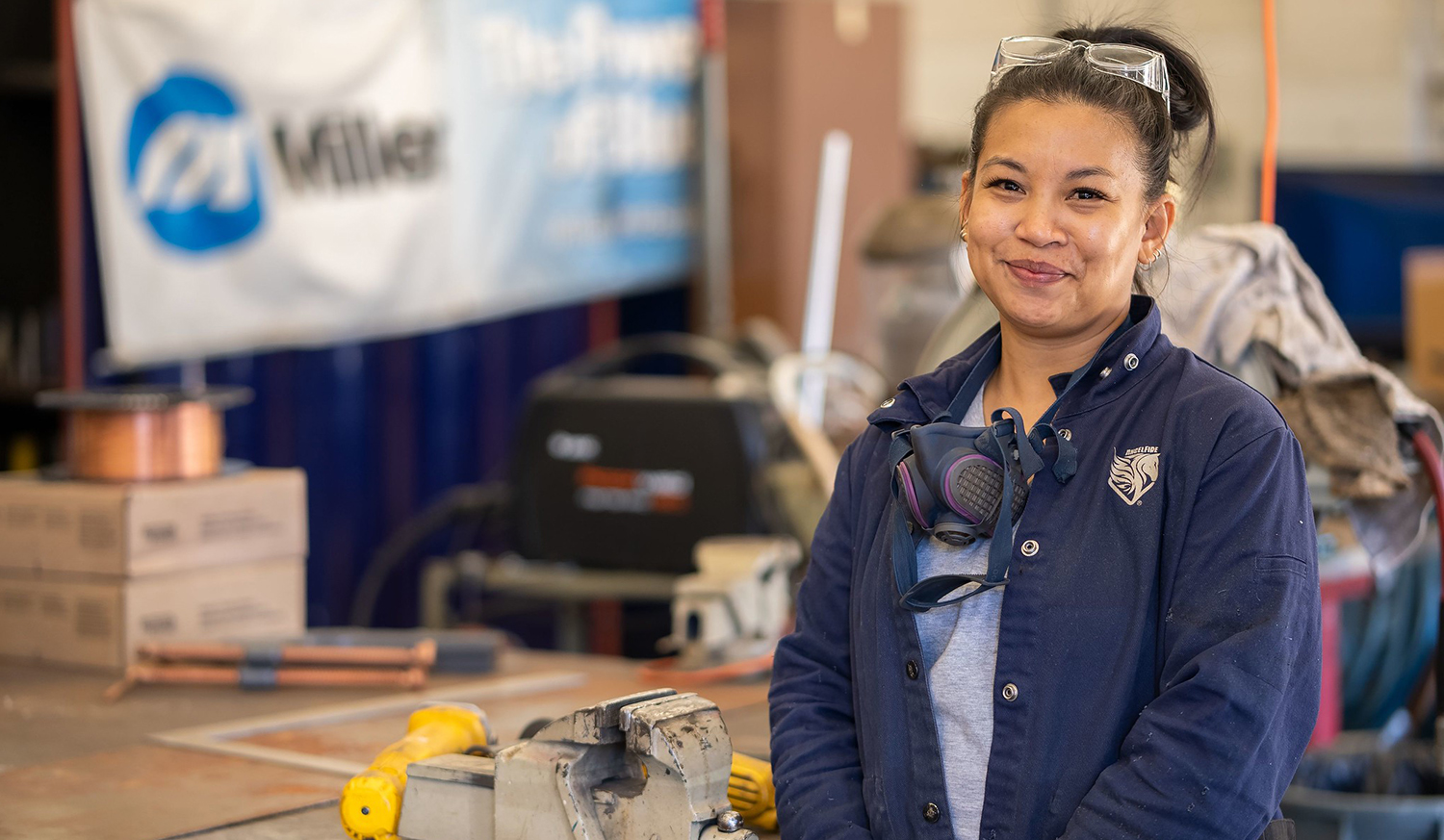 Samantha Marasigan in a workshop stands next to a vice and wears a blue jacket, safety goggles on her head and a facemask around her neck