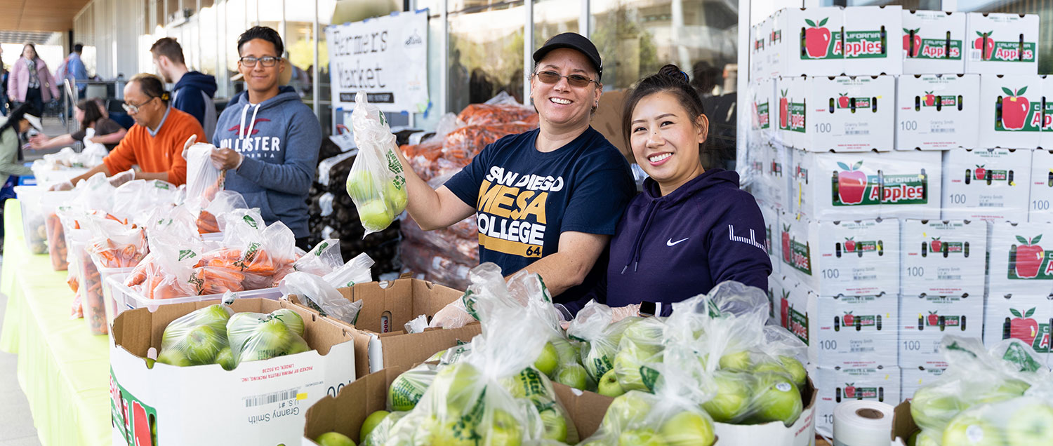 four workers are at the free farmers market ready to distribute food to students. They standing behind long folding tables with apples and carrots for people to take.