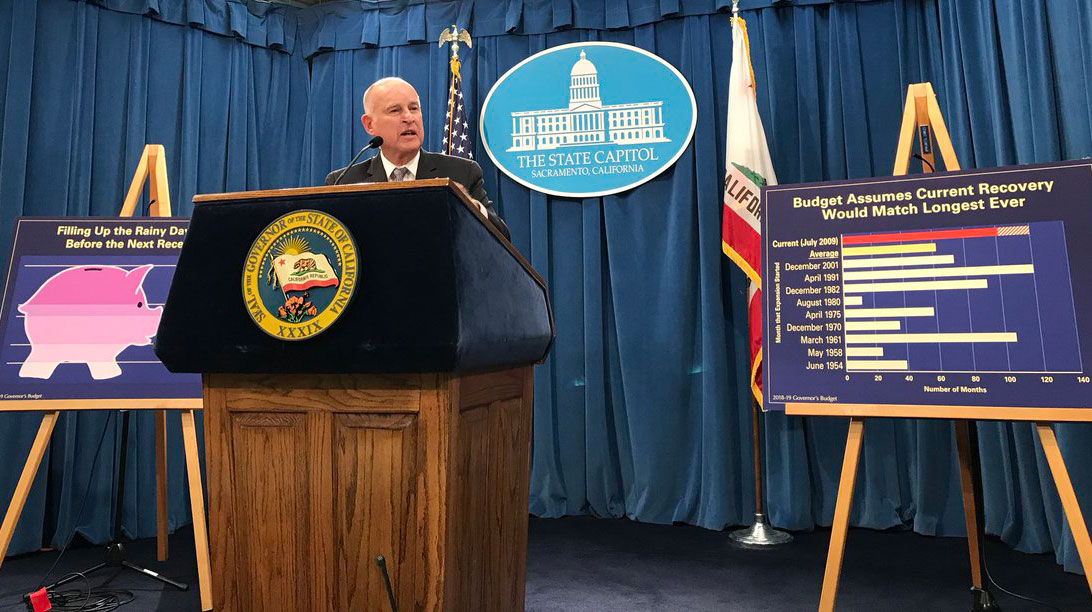 Governor Jerry Brown stands at a podium at a budget press conference