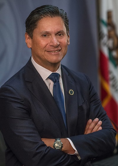 Portrait photo of Eloy Ortiz Oakley wearing a blue suit, seated in front of a California flag