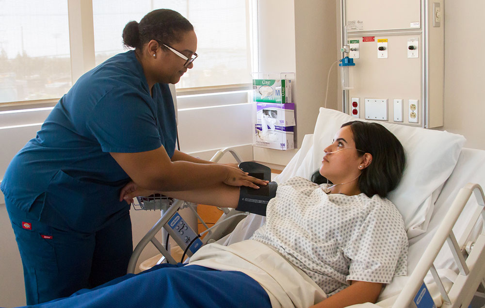 A healthcare student takes the blood pressure of another student who is in a hospital bed.