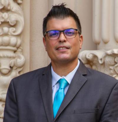 Carlos O. Cortez portrait photo. He is wearing a gray suit and teal tie