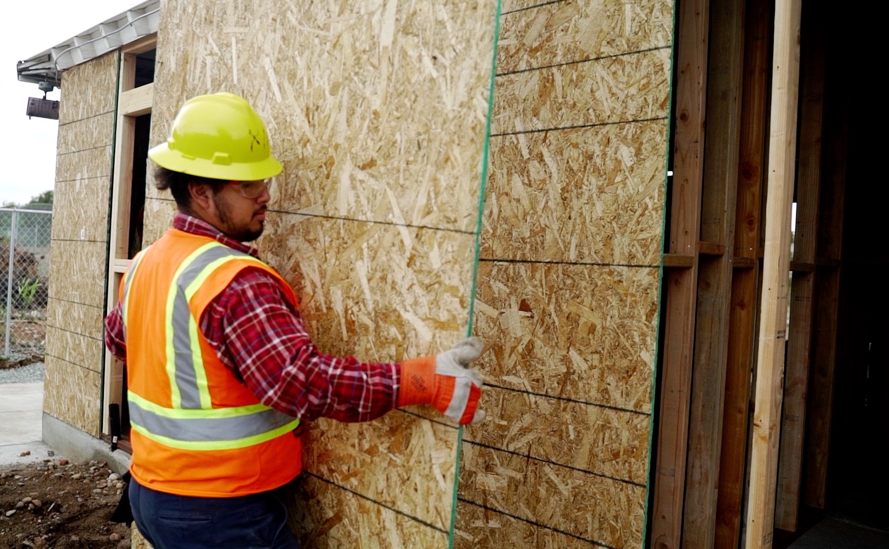 A construction student carries some plywood at a construction site.