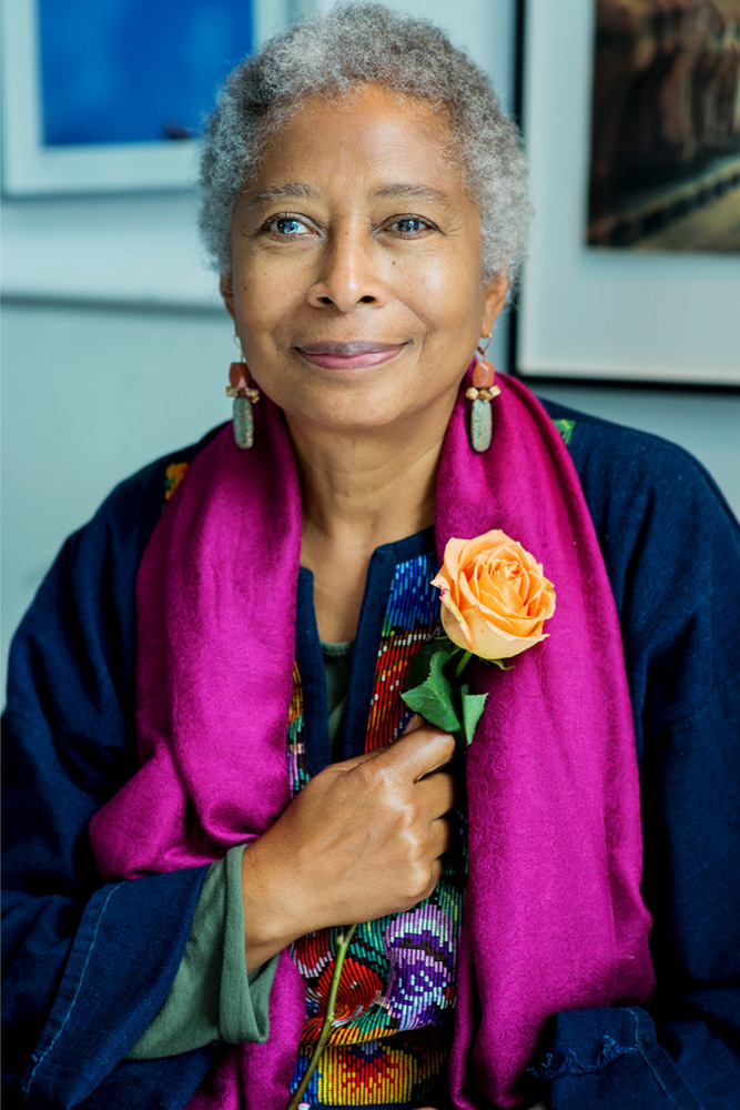 A portrait photo Alice Walker wearing a bright pink scarf and holding an orange rose 