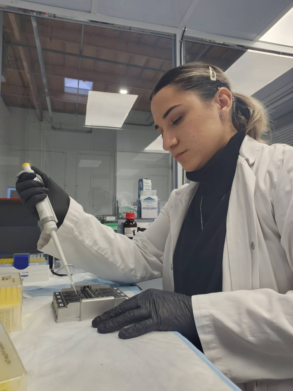 Andrea fills vials with a syringe at a work station. She is wearing a white coat and black rubber gloves.