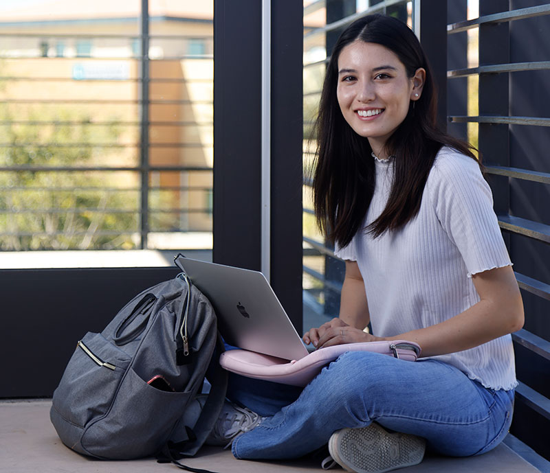 Emily Smith sits on the floor next to her gray backpack and works on her laptop