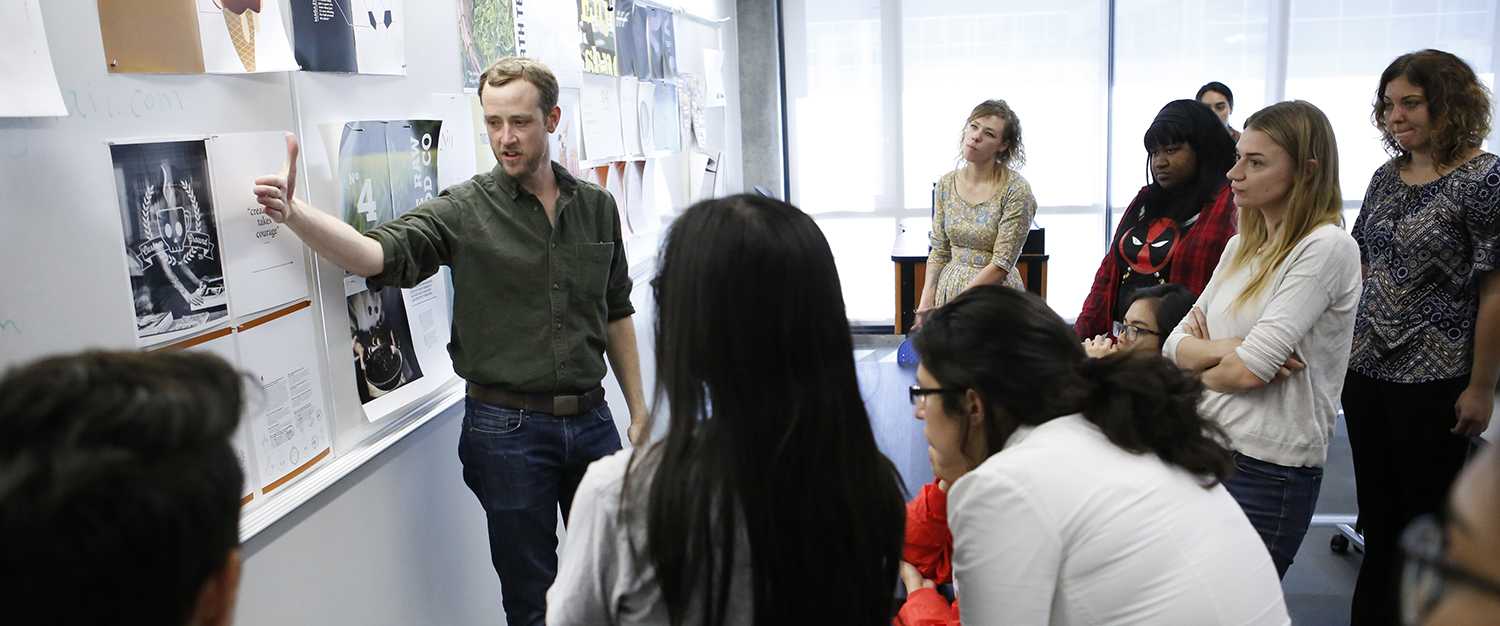 A teacher points to information on a white board as several students gather in a semi circle to read the board