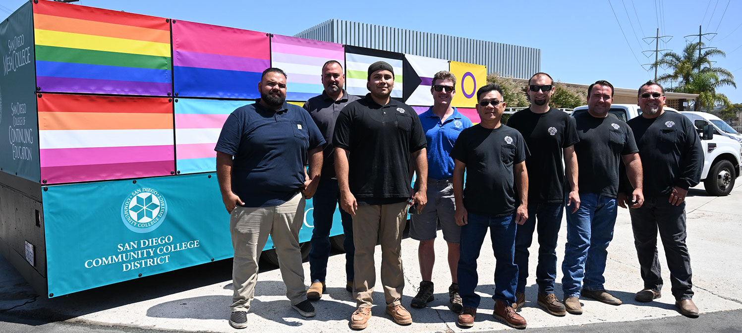 Eight men in dark t-shirts, jeans and work boots stand in front of the pride float which is covered in many different pride flags.