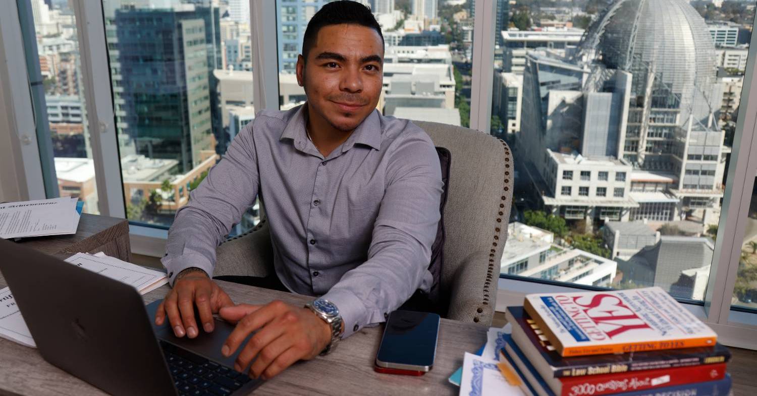 Frank Hernandez sits a his desk in his office. He is typing on his laptop. There is a view of the San Diego skyline outside of his office window