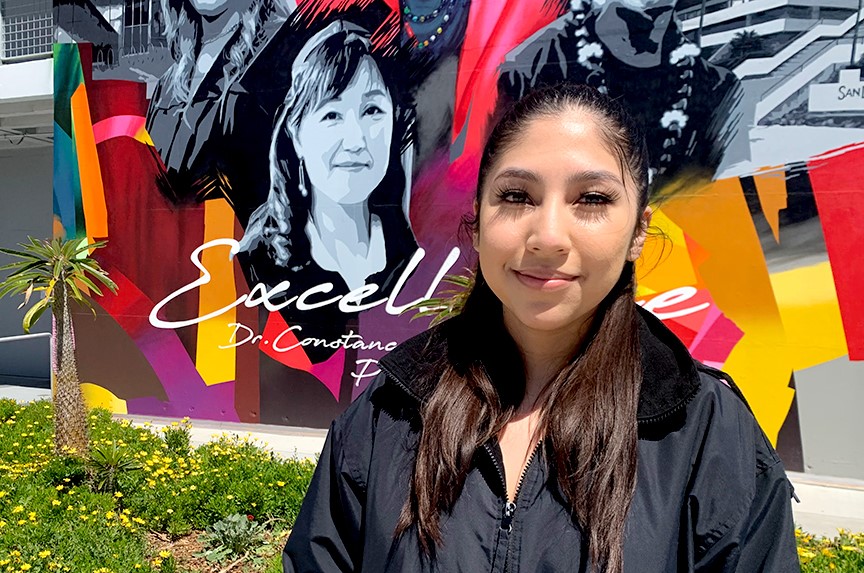 Jeannette Mayo Gallegos wears a black windbreaker jacket and is standing in front of a colorful mural at City College