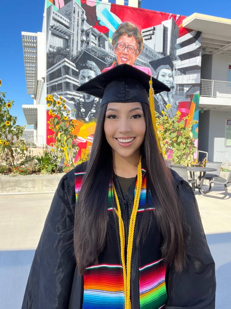 Jeannette Mayo Gallegos wears a black graduation cap and gown and is standing in front of a colorful mural at City College
