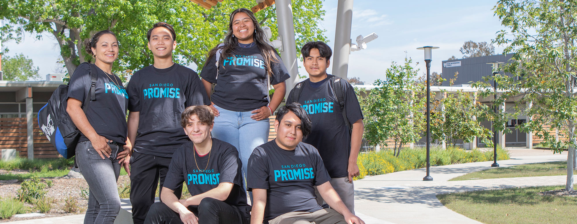 Six students wearing promise t shirts are outside on a bench in the Mesa Quad with greenery in the background.