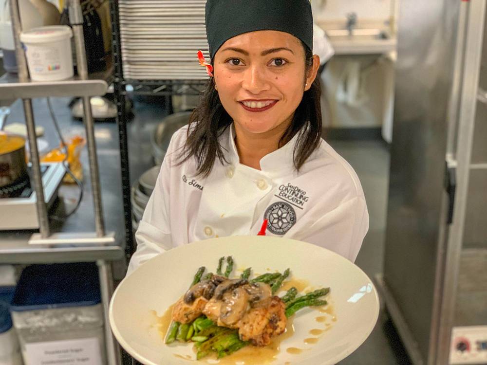 A chef in a kitchen wearing a chef's had and smock holds up a plate of food