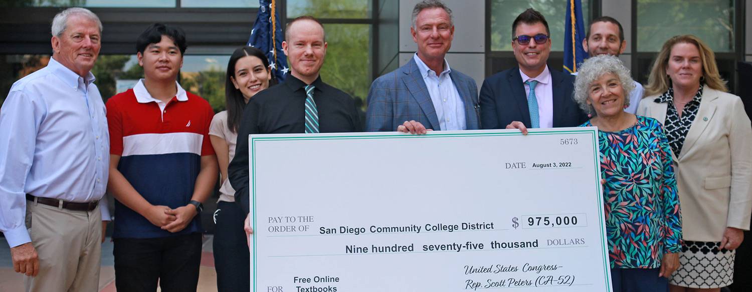 Rep. Scott Peters is in the center of the photo wearing a gray, blue blazer, a light blue button down shirt and tan pants. He's holding a giant check for 975,000 dollars and is surrounded by 6 district officals and 2 students.