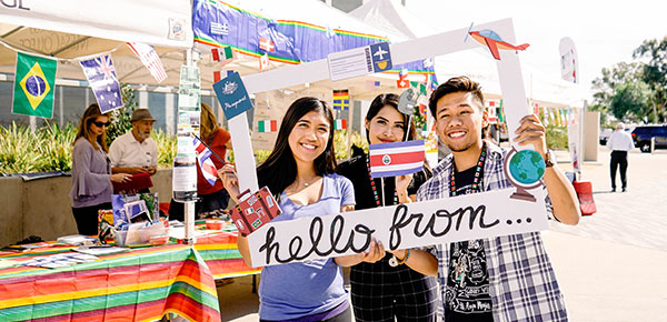 Three study abroad students hold up a giant cardboard picture frame that reads hello from ...