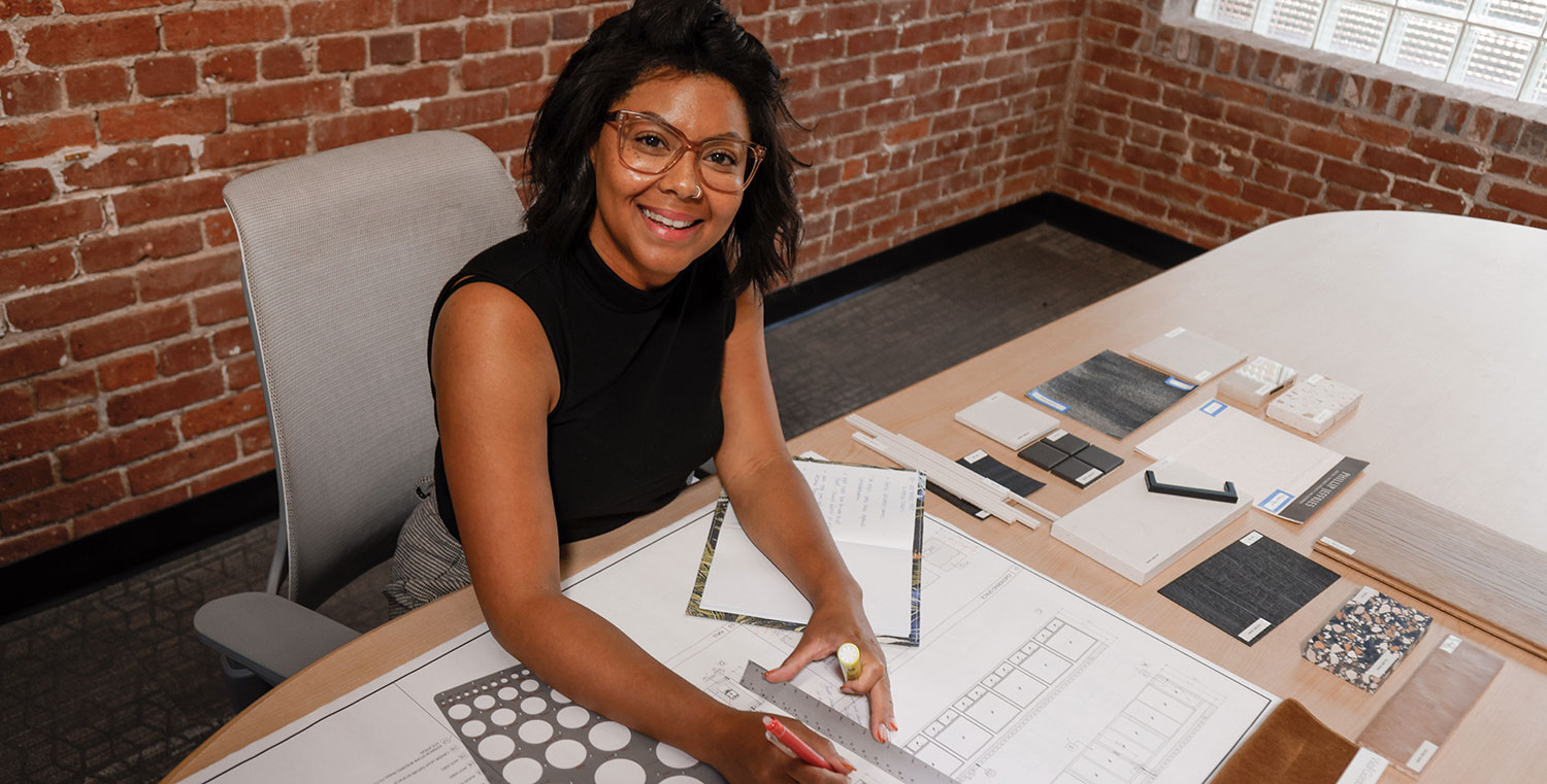 A graduate sits in here office at her desk with designs and drawing tools.