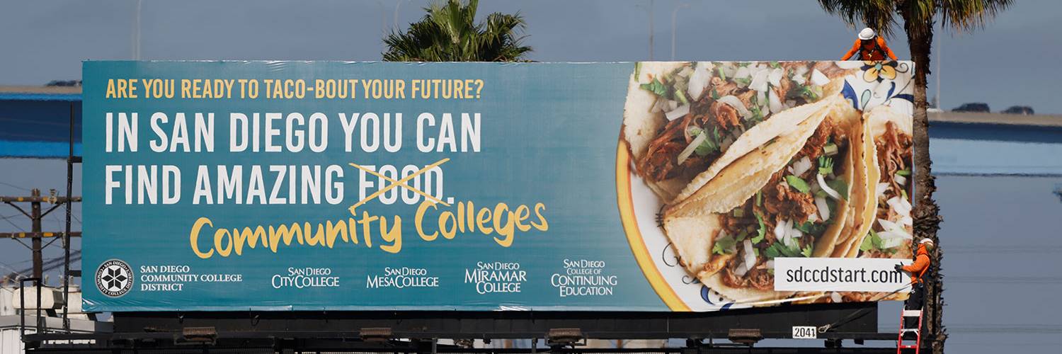 Marketing Billboard Tacos Workers install a billboard. The billboard shows a plate of taco. Text reads Are you ready to taco bout your future In San Diego you can find amazing food which is crossed out and replaced with community colleges.