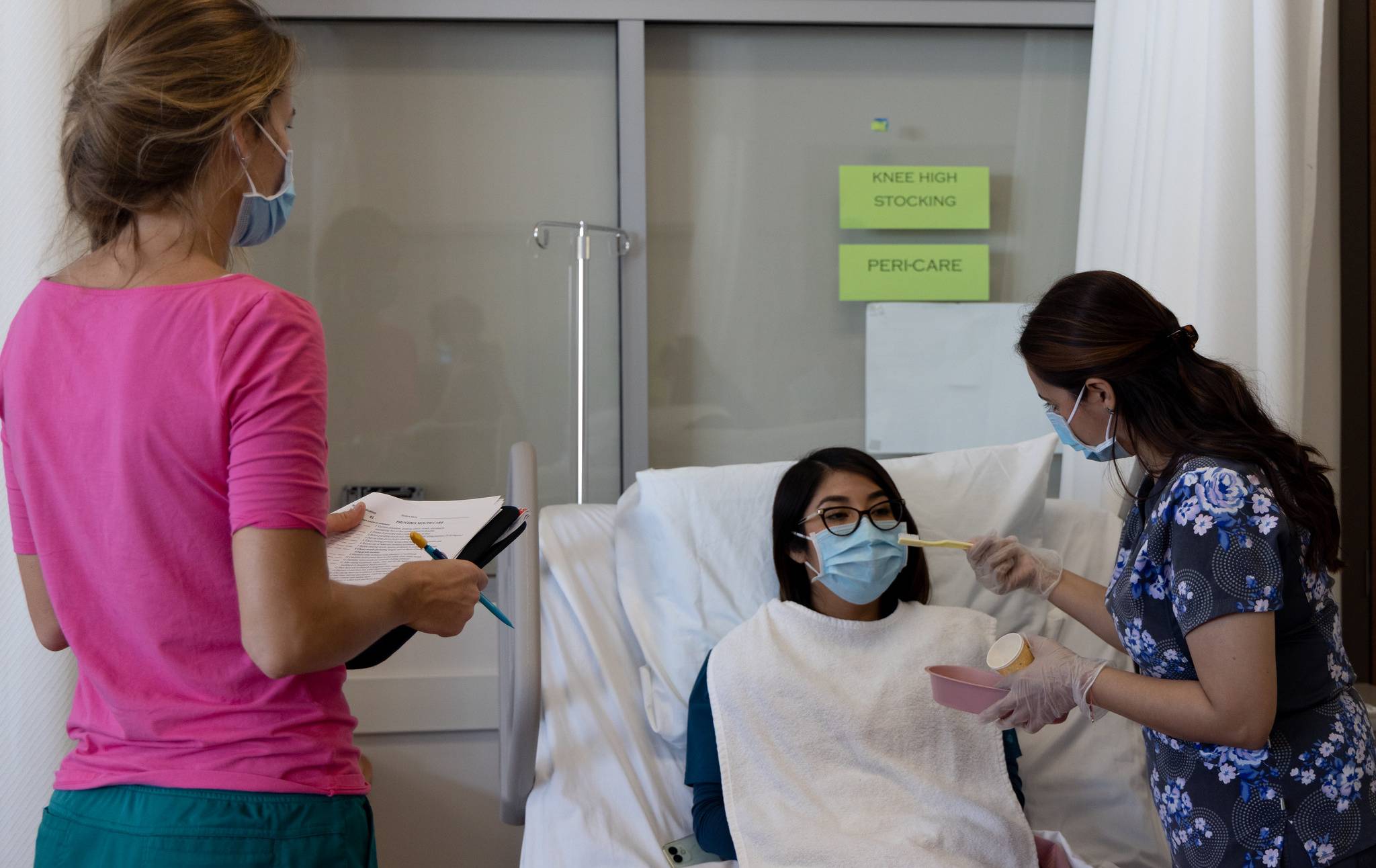 A nursing student holds a toothbush for another student in a hospital bed acting as a patient. An instructor looks on