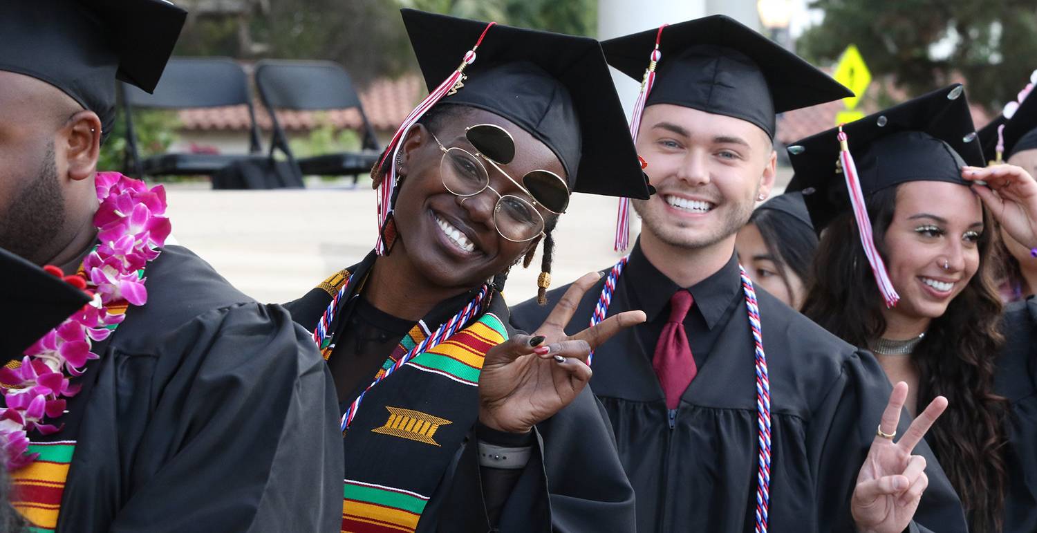 Two students wearing black caps and gowns give peace signs as they line up to walk the stage at commencement