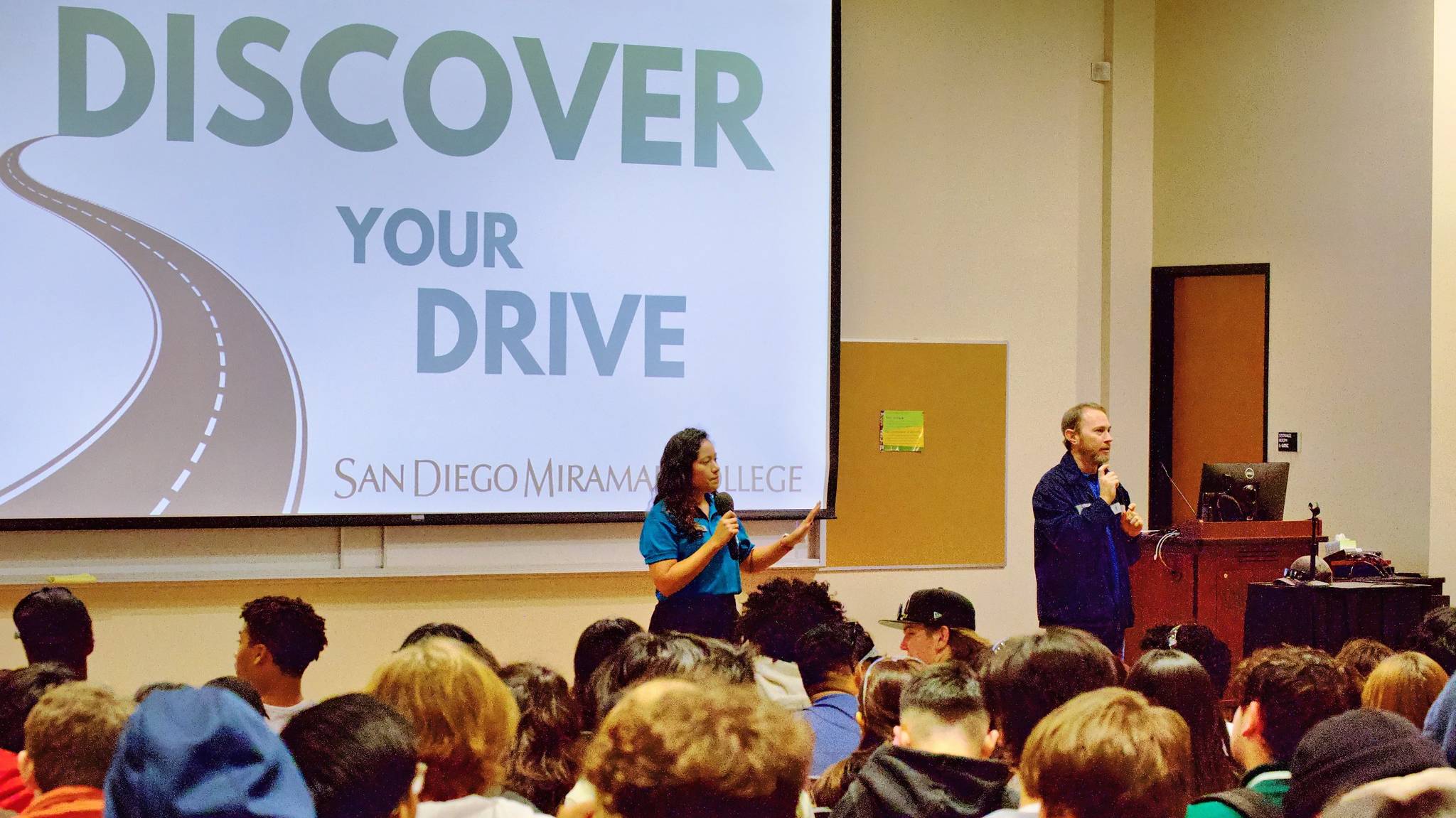 A woman in a teal polo shirt speak to an auditorium of high school students.