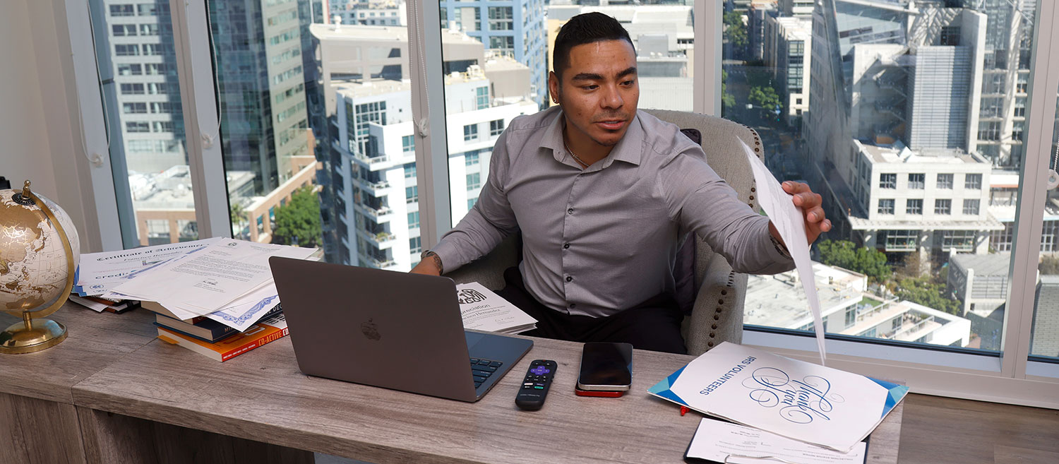 Frank Hernandez sits at his desk in his office. A laptop is on his desk. He is looking over papers on his desk. There is a view of the San Diego skyline outside of his office window