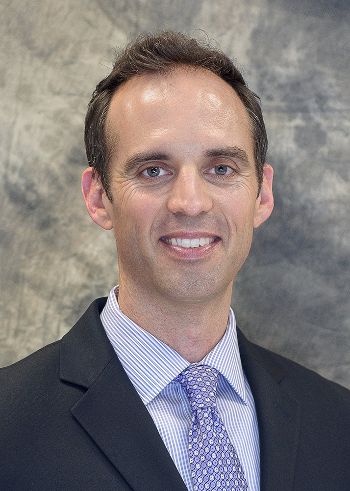 Portrait photo of Gregory Smith wearing a black suite and a blue shirt and tie