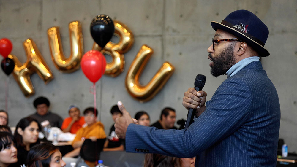 A man speaks to an audience. Gold balloons on the wall spell Hubu