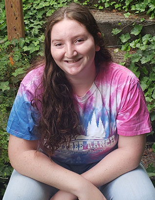 Kimberly Kritzer is sitting on stairs outside surrounded by greenery. She is wearing a tie dye t shirt