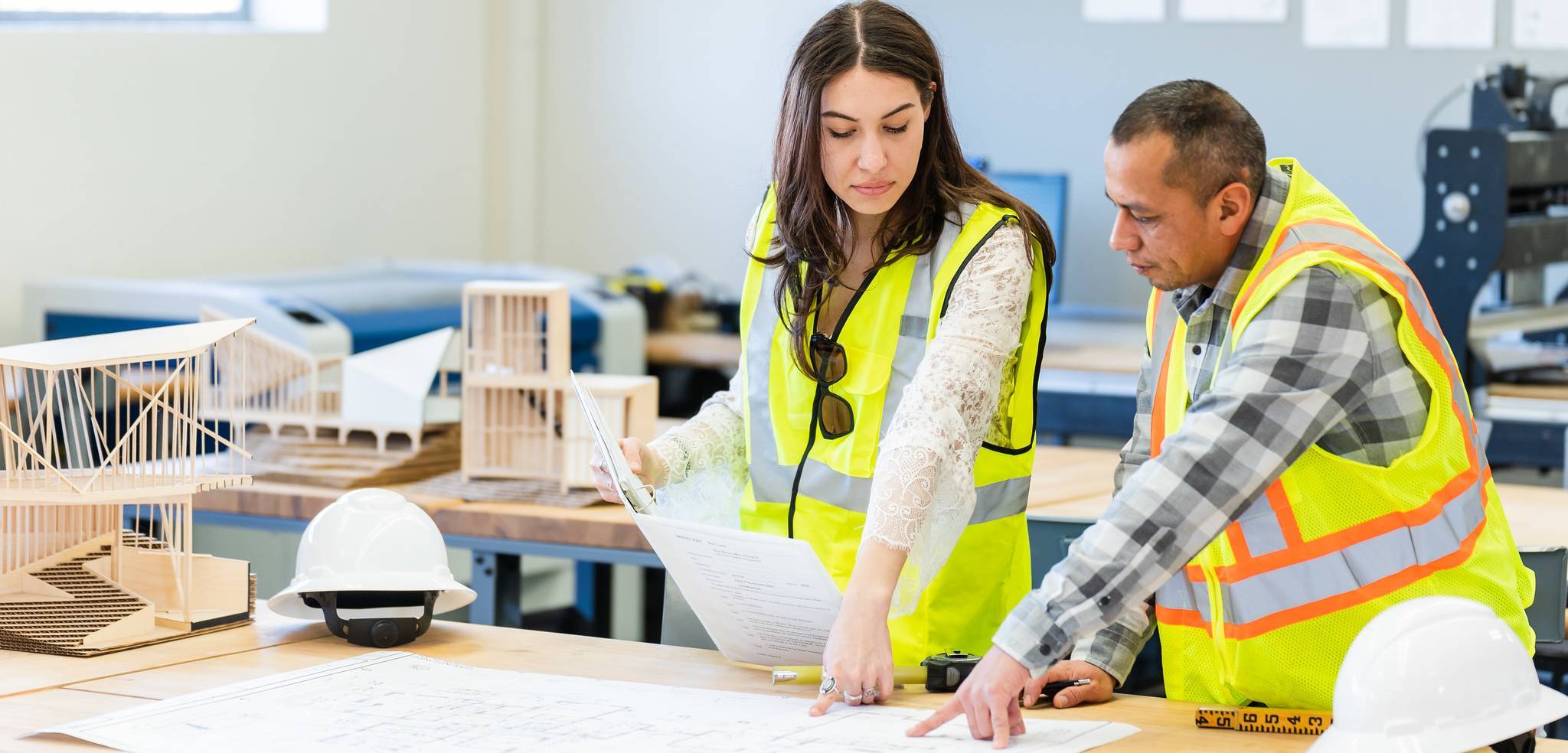 Mesa Architecture A man and a woman are looking at architecture plans at a work table. They are wearing yellow reflective vests and two white hard hats are on the table.