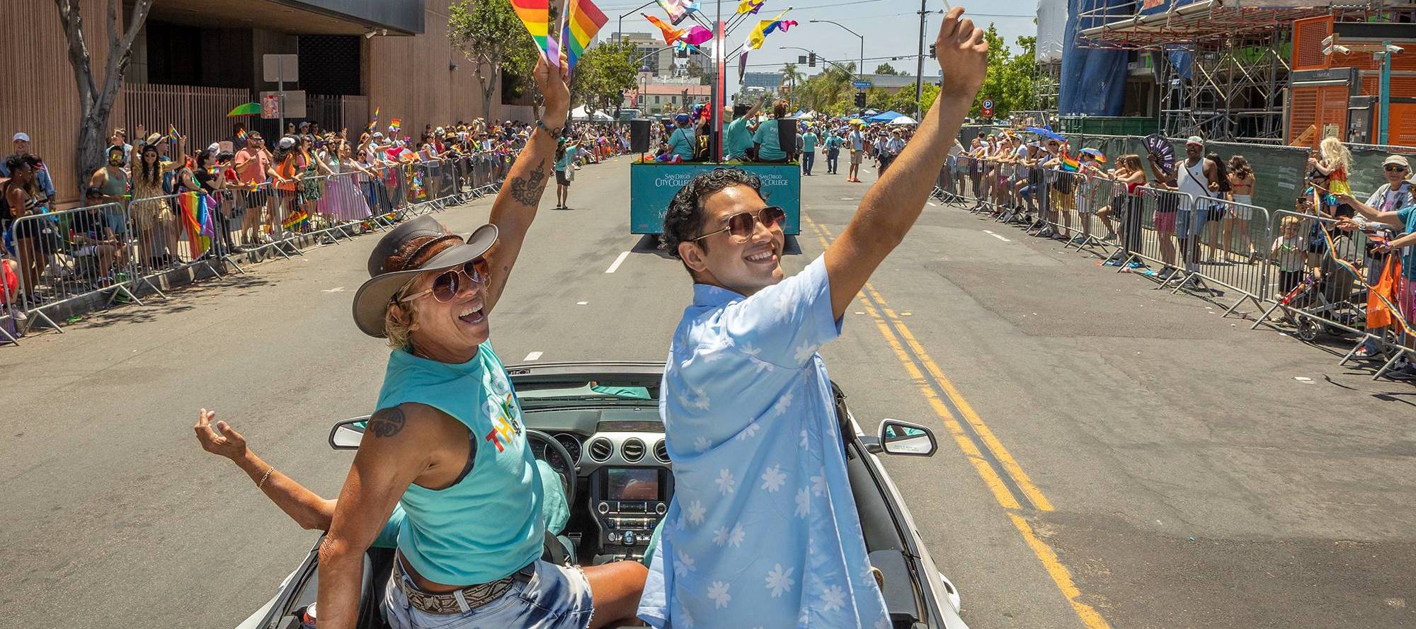 Two people sit on top of the back seat of a convertible car and wave during the pride parade