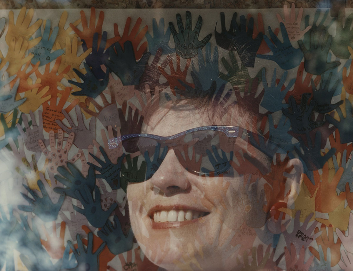 A woman wearing sunglasses looks through a window at colorful hand print artwork. The artwork is not visible, but reflected in the window.