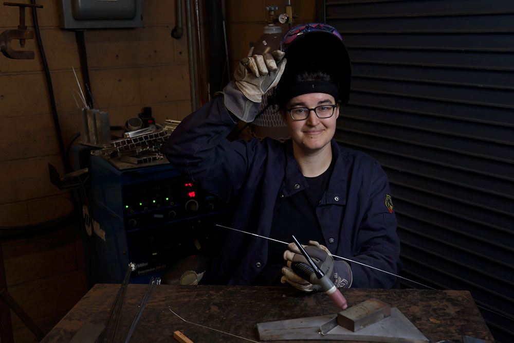 Sarah McKee wears welding gear including thick gloves, a welding mask and a protective jacket. She is holding a welding torch