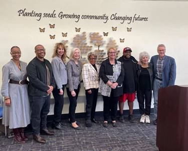 Nine people take a photo in front of the wall sign at the center. The wall has a tree made of brass leaves that is surrounded by plastic butterflies. Text reads planting seeds growing community changing futures