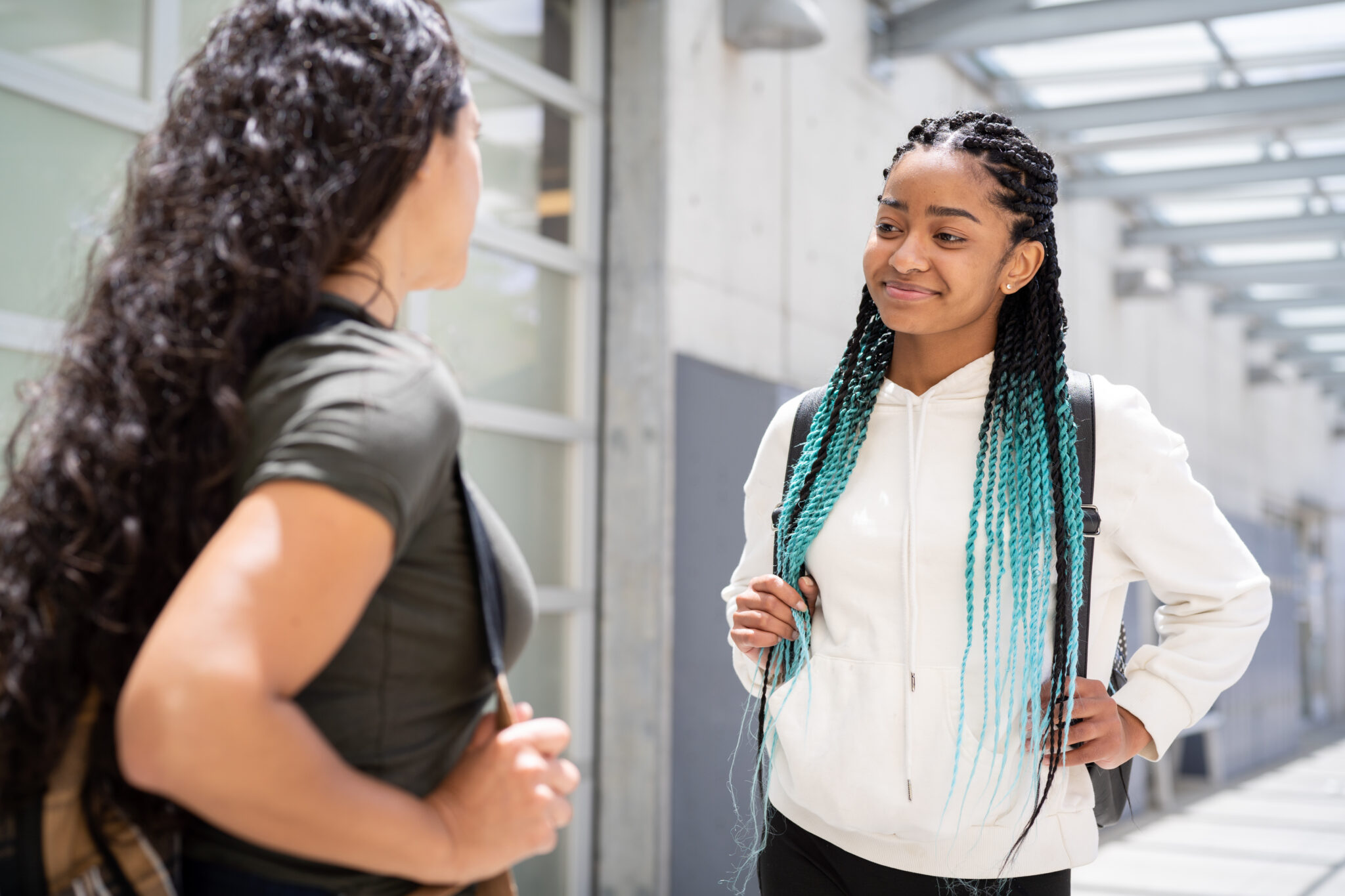 A student wearing a white button down shirt, is wearing her hair in braids with teal tips, speaks to another student on campus