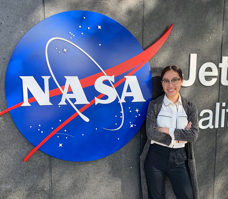 Katya stands next to a large blue sign of the NASA logo