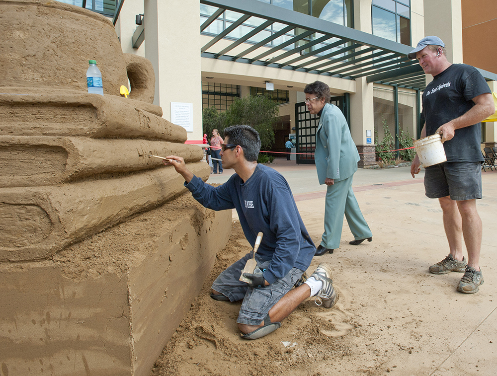 A sand sculpture of a stack of books one man is working on the sculpture and two people are watching.