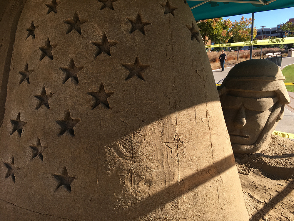 A sand sculpture for veterans day shows a soldiers head wearing a helmet. A wall of sand has stars carved into it.
